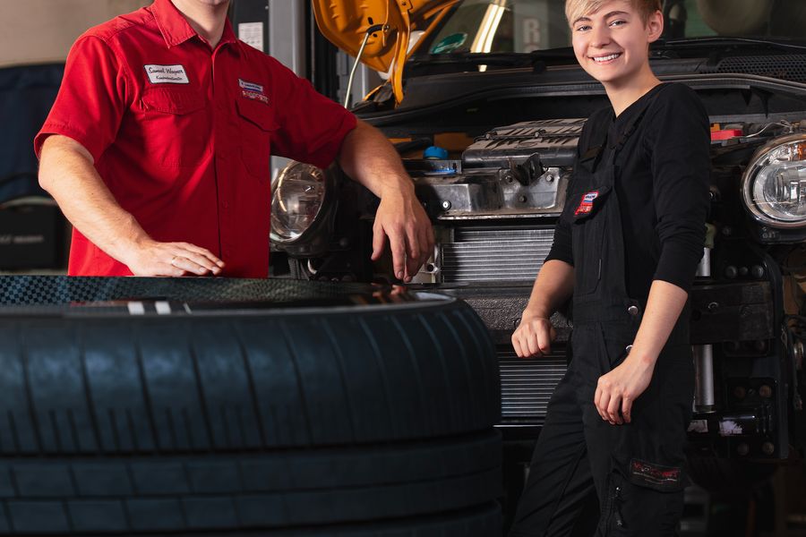 Kfz-Meister und Auszubildende stehen lächelnd neben einem Fahrzeug mit geöffneter Motorhaube in der Werkstatt