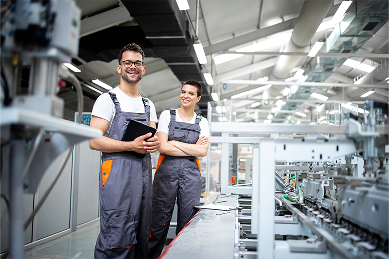 Zwei Fachkräfte in Arbeitskleidung stehen in einer Produktionshalle und blicken in die Kamera