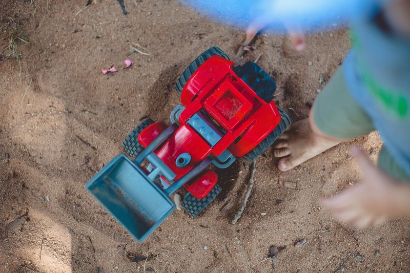 Ein roter Spielzuegbagger in einem Sandkasten von oben fotografiert. Daneben steht ein Kind, es ist barfuss und man kann nur seine Füße sehen.