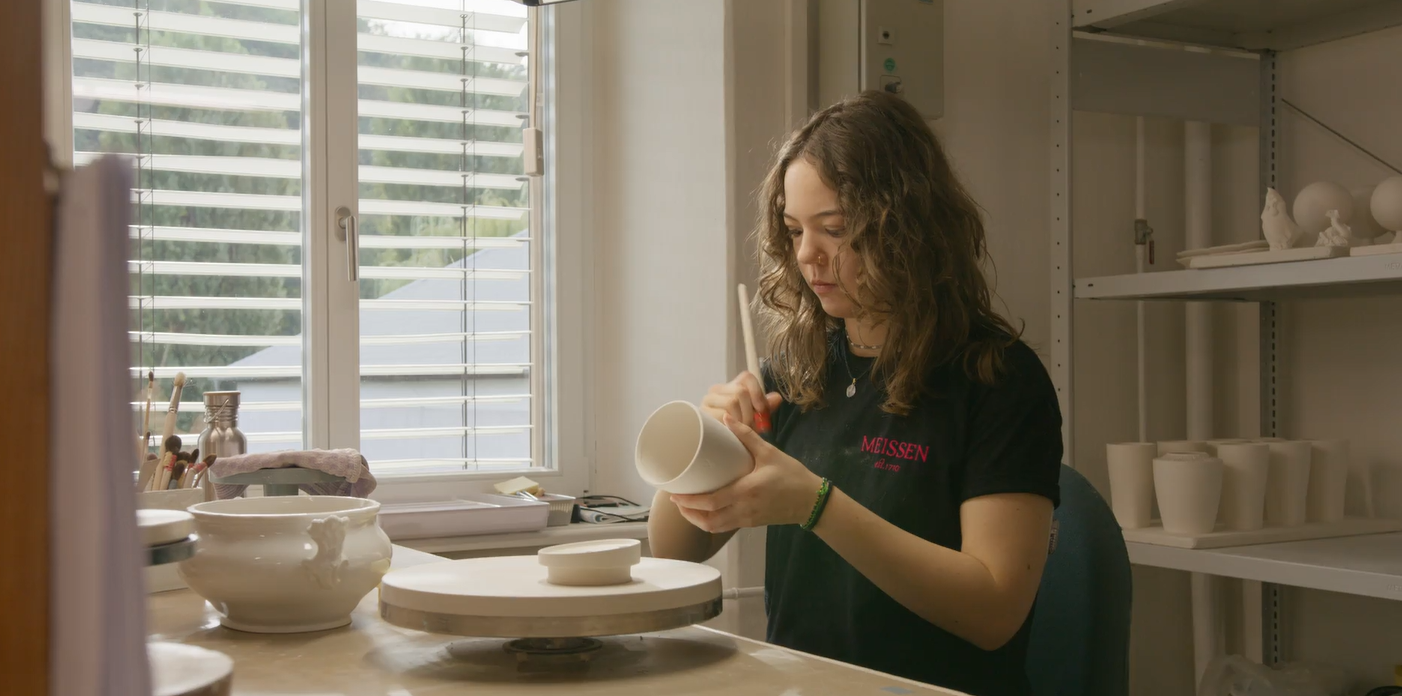 Porzellanmalerin bemalt in einer Werkstatt eine Vase, im Regal weiteres Weißporzellan