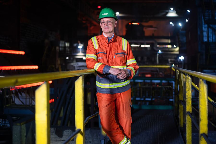 Ein Mann in einem orangefarbenen Schutzanzug und grünem Helm steht in einer Stahlfabrik. Er lehnt an einem Geländer, umgeben von industrieller Ausrüstung und rot glühendem Stahl im Hintergrund.