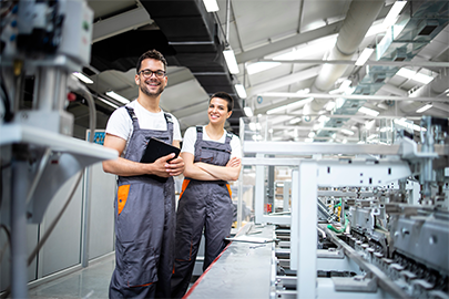 Zwei Fachkräfte in Arbeitskleidung stehen in einer Produktionshalle und blicken in die Kamera