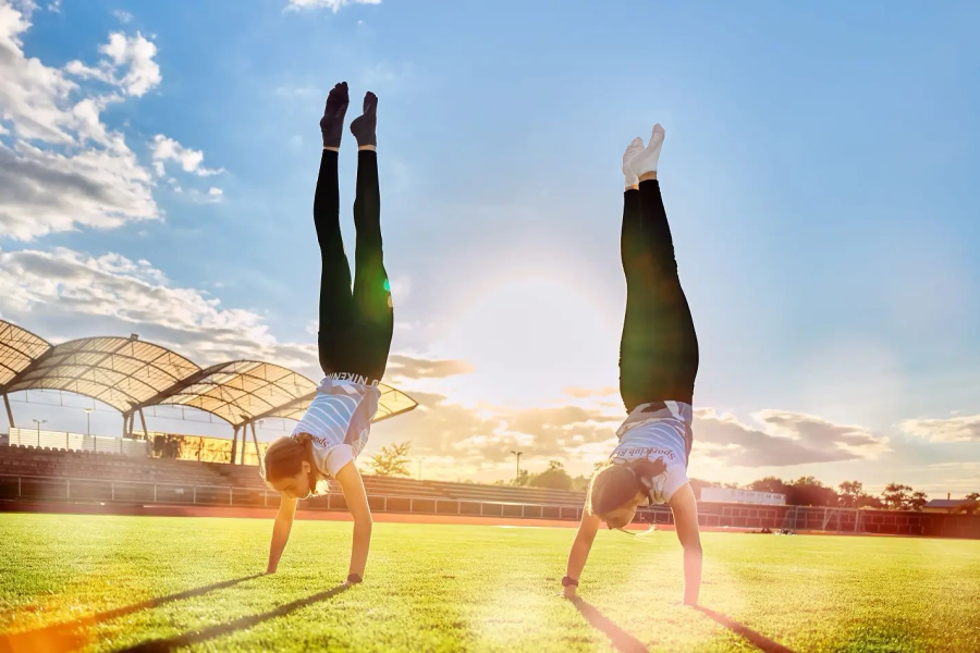 Zwei Sportlerinnen machen bei Sonnenschein einen Handstand auf einem Sportplatz
